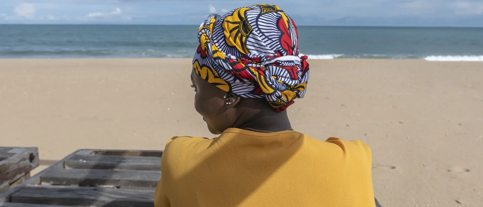 A woman wearing a colorful headwrap sits by the beach, facing the ocean, with a sandy shore in the background.