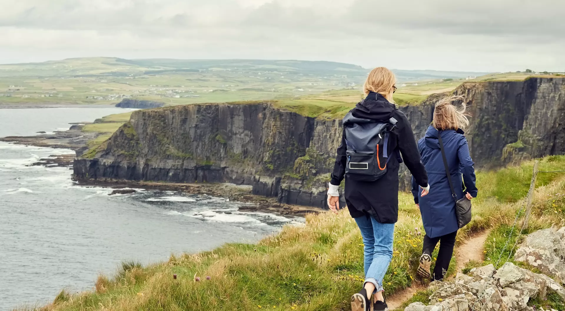Two people walk along a grassy path overlooking a coastal cliff and ocean under a cloudy sky.