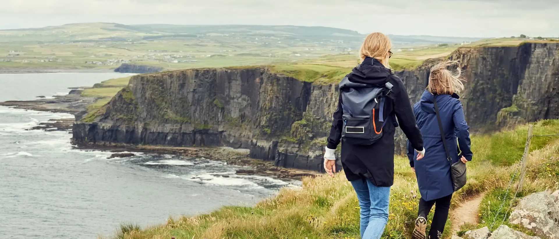 Two people walk along a grassy path overlooking a coastal cliff and ocean under a cloudy sky.