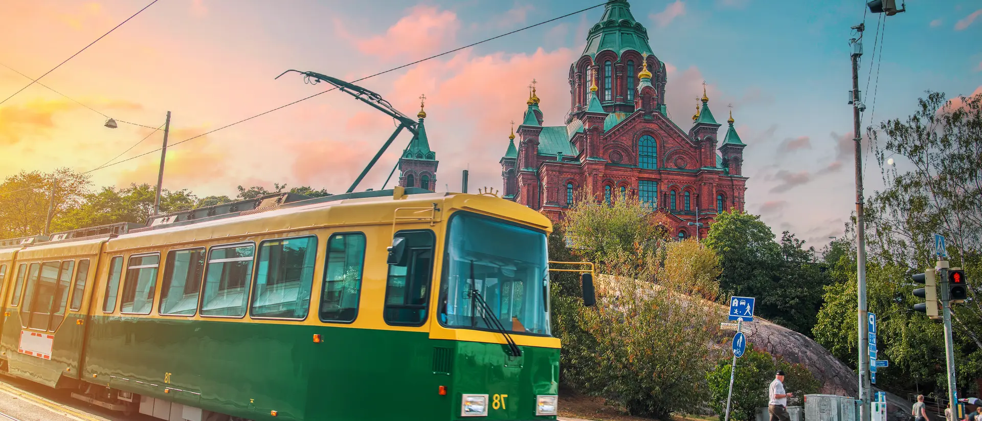 A green and yellow tram travels on a street with a historic building in the background at sunset.