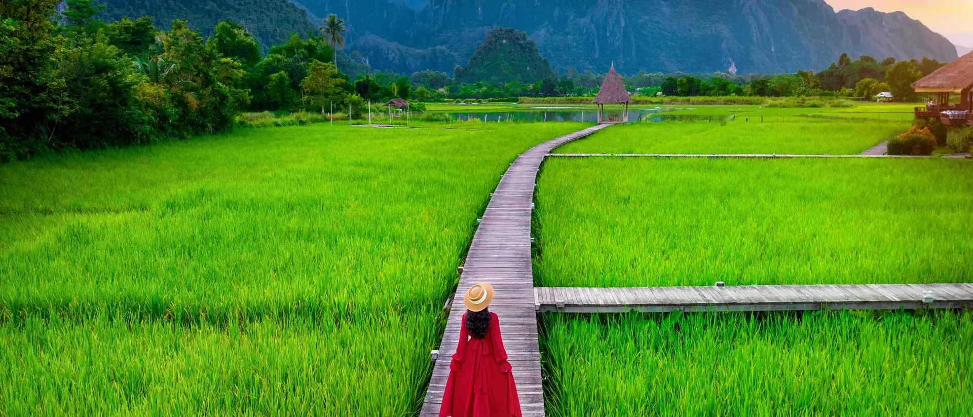 A woman in a red dress walks along a wooden path through vibrant green rice fields with mountains in the background.