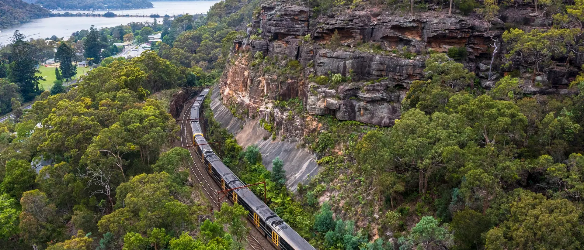 A train curves along a track beside a rocky cliff and lush greenery, with a lake visible in the background.