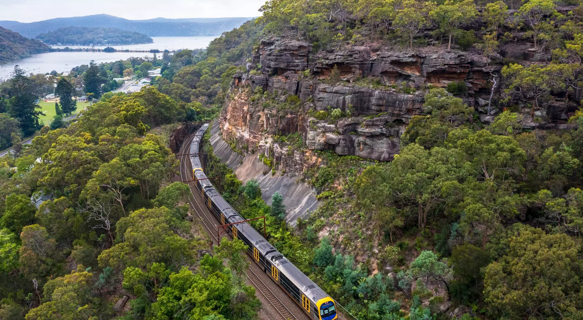 A train curves along a track beside a rocky cliff and lush greenery, with a lake visible in the background.