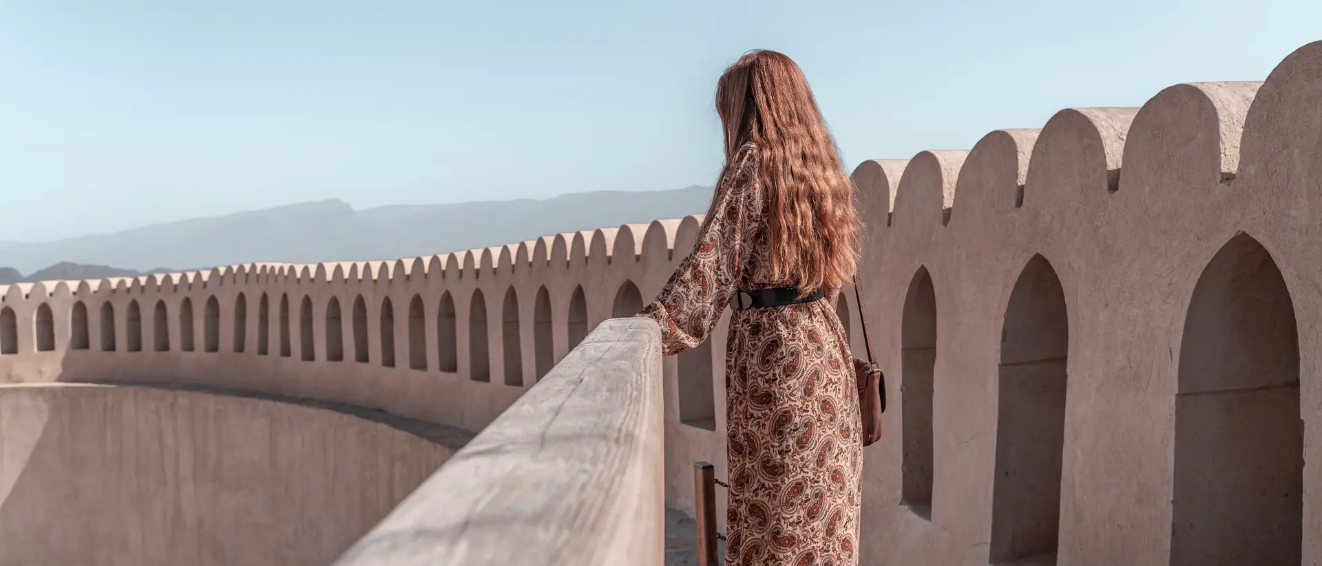 A woman in a patterned dress stands on a balcony, gazing at distant mountains under a clear blue sky.