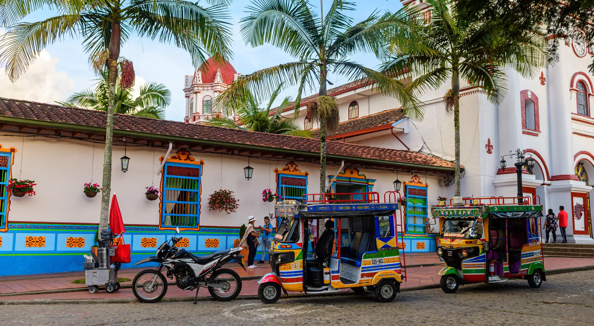 Colorful tuk-tuks and a motorcycle park beside a vibrant colonial building with palm trees and decorative flowers.