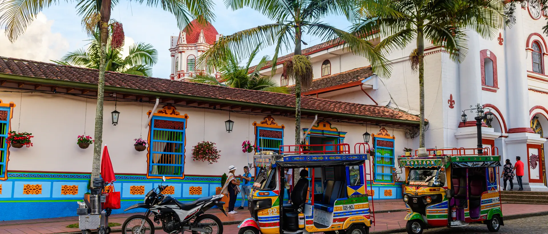 Colorful tuk-tuks and a motorcycle park beside a vibrant colonial building with palm trees and decorative flowers.