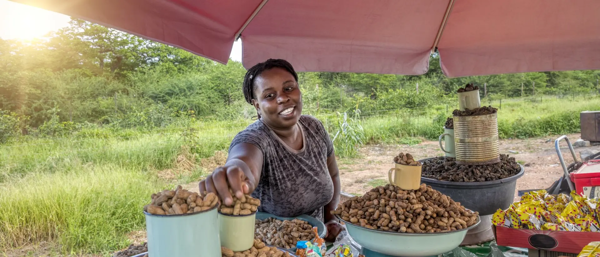 A smiling vendor at a market offers a cup of peanuts, surrounded by various snacks under a shaded tent.