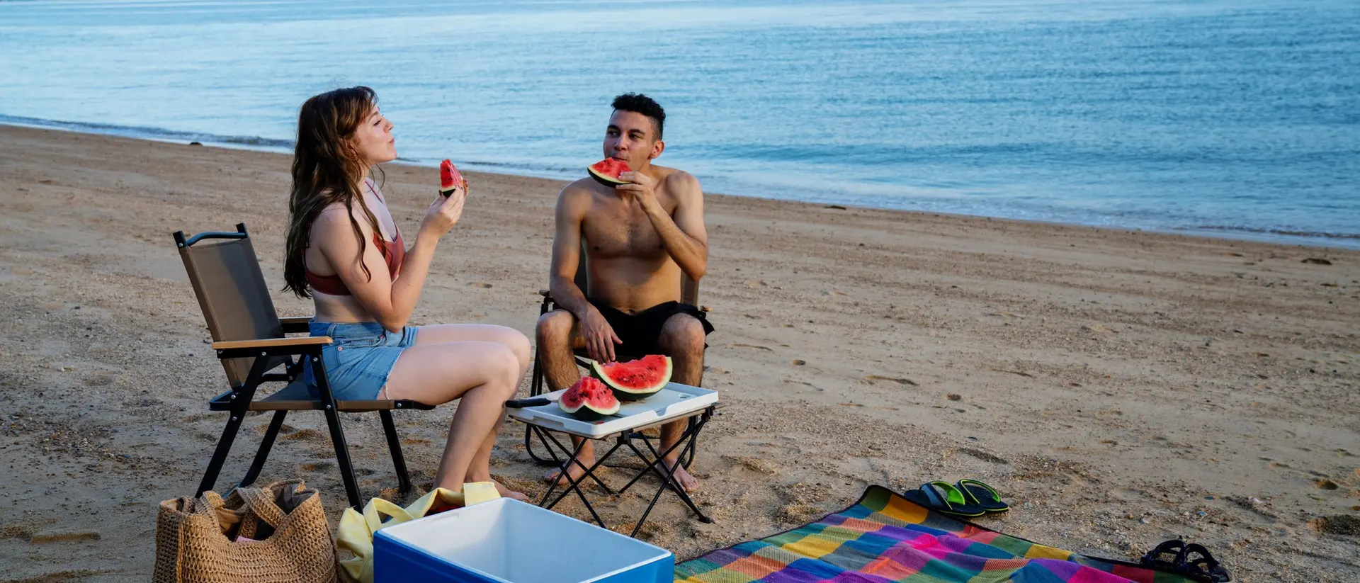 Two people enjoying watermelon on a beach, sitting on a colorful blanket with a cooler nearby and ocean in the background.