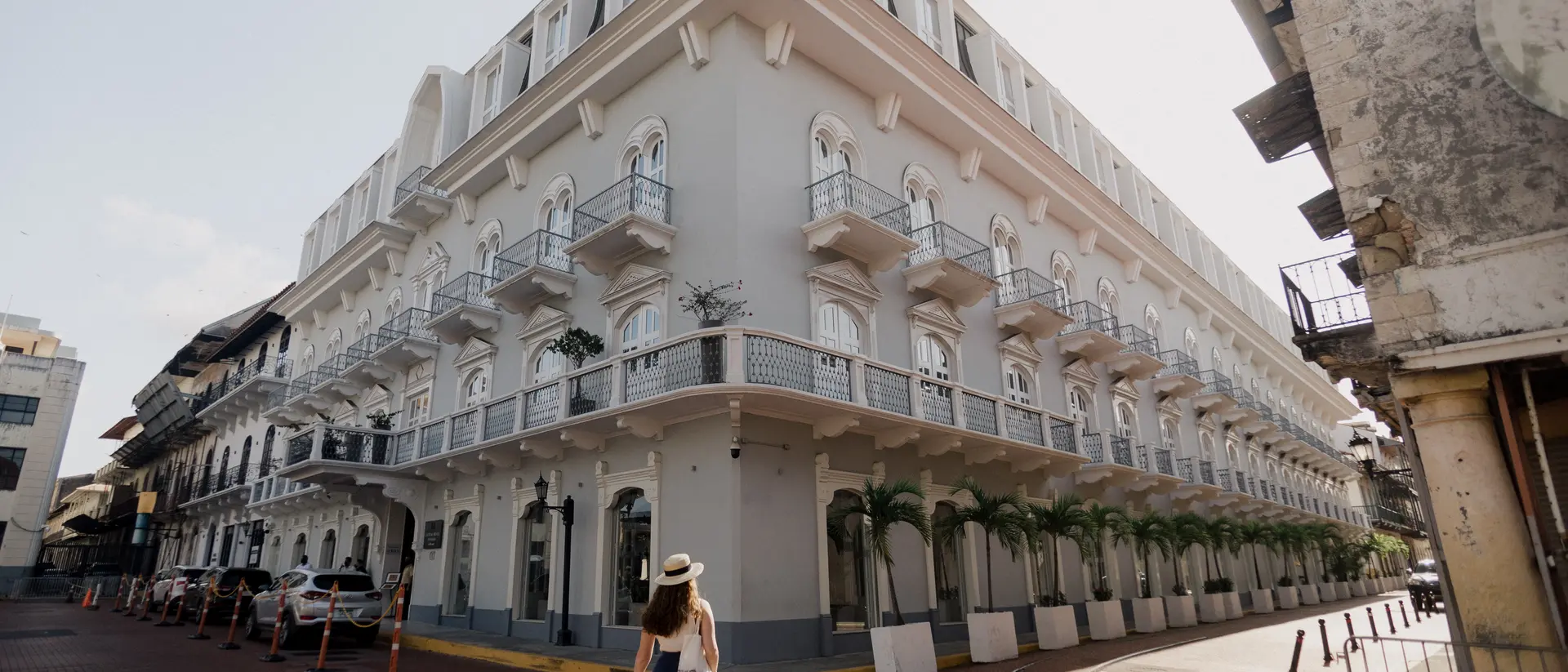 A woman walks along a cobblestone street beside a large, elegant white building with balconies and palm trees.