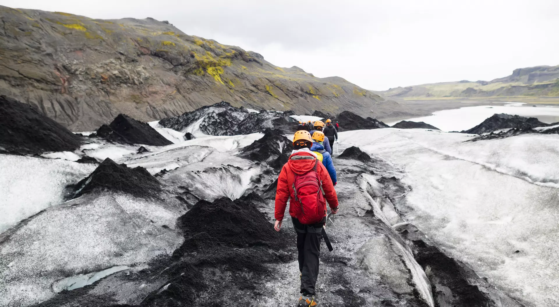 A group of hikers wearing helmets walks on a glacier with rocky terrain and a cloudy sky in the background.