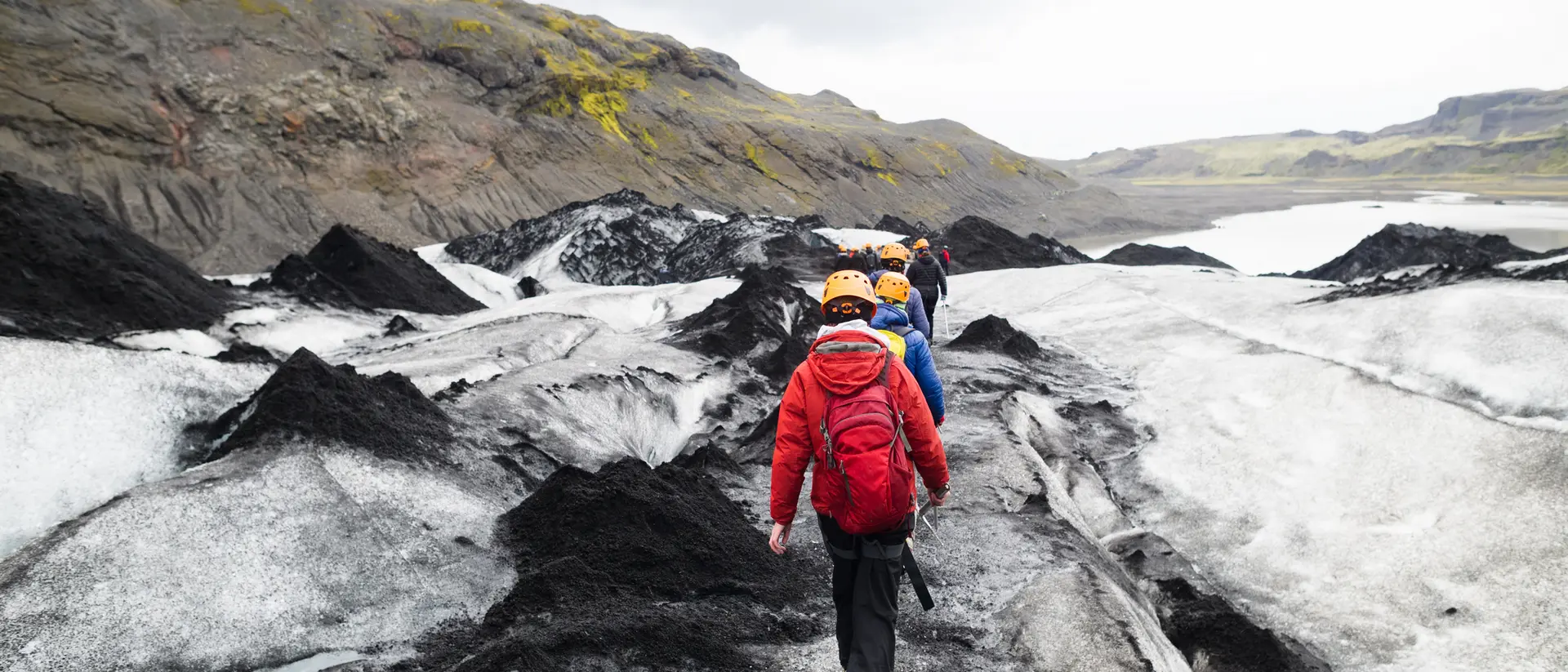 A group of hikers wearing helmets walks on a glacier with rocky terrain and a cloudy sky in the background.