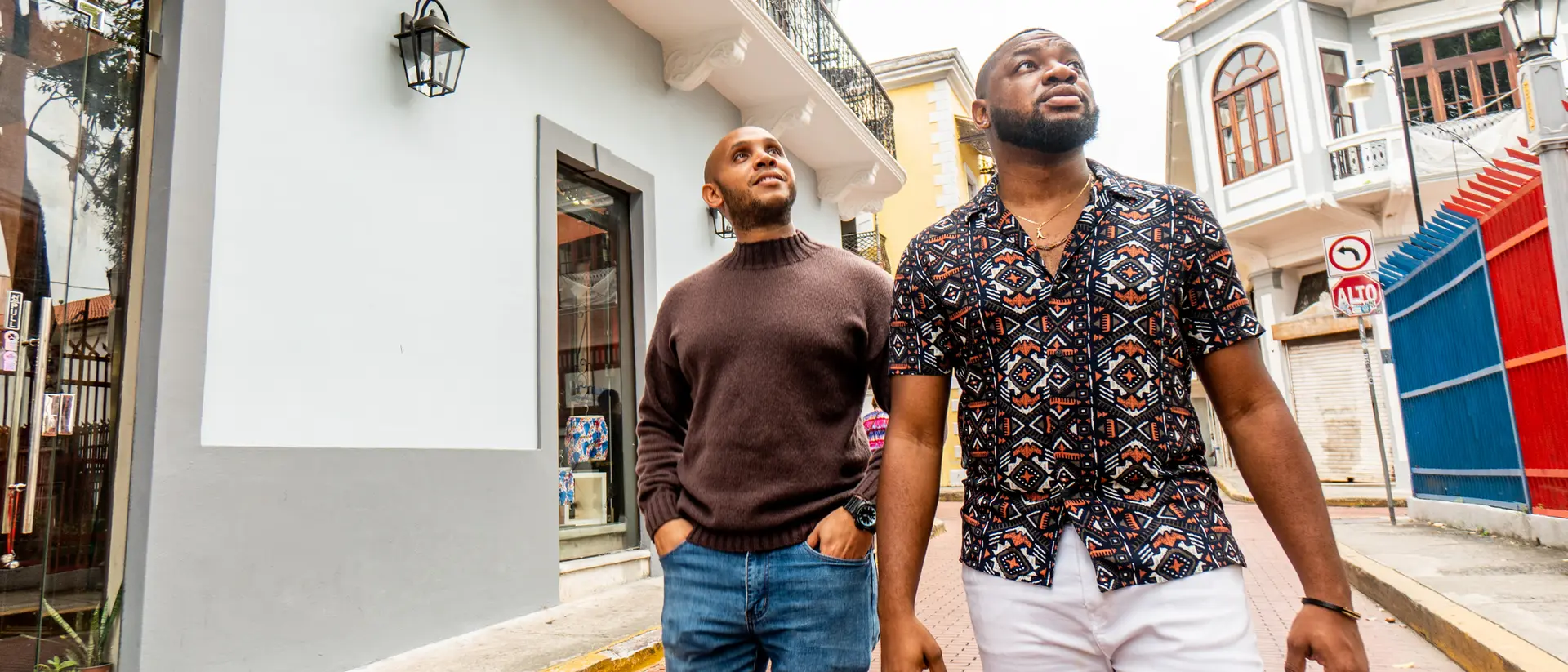 Two men walking down a vibrant street, with colorful buildings and shops in the background.