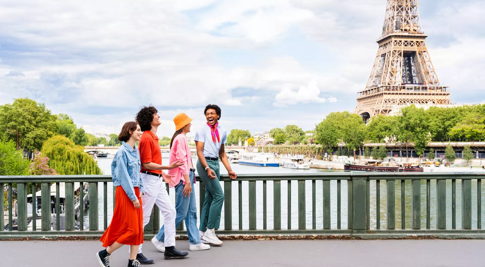 Four friends walk along a bridge with the Eiffel Tower in the background, enjoying a sunny day by the river.