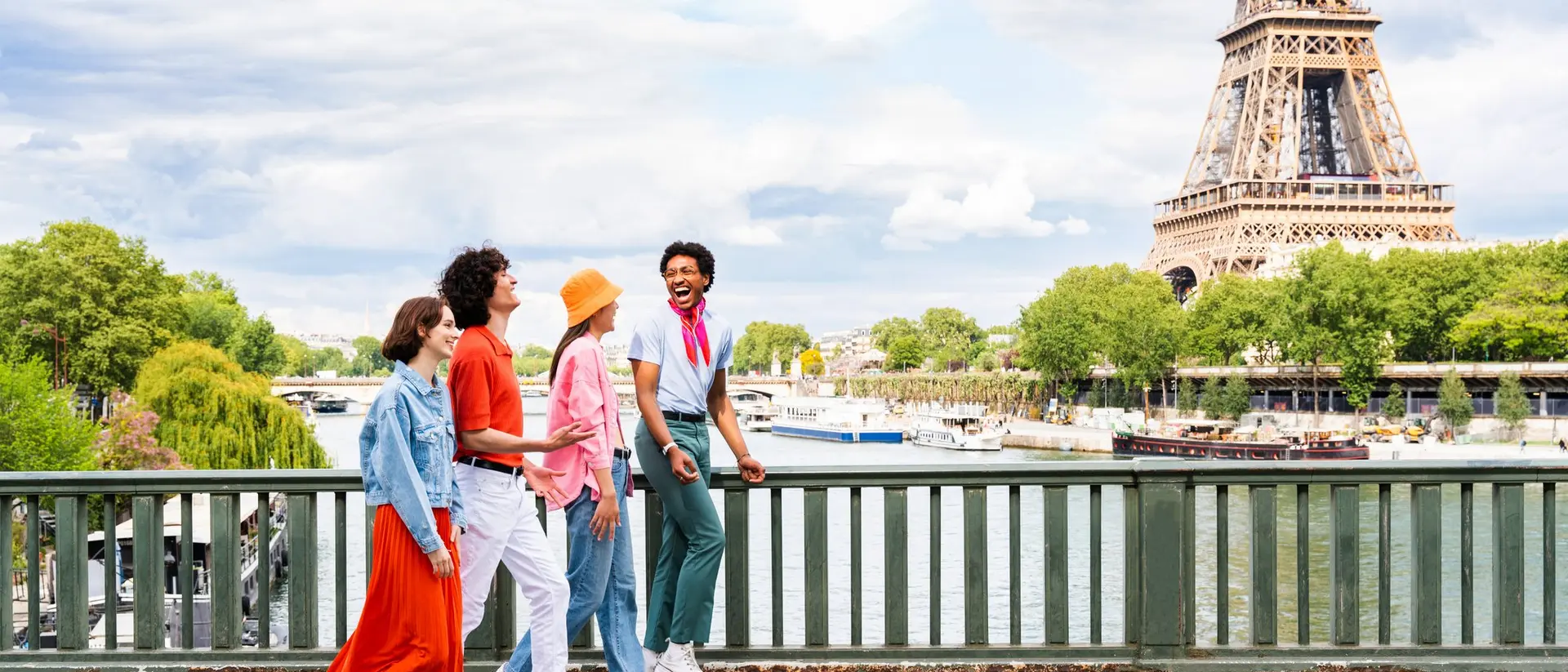 Four friends walk along a bridge with the Eiffel Tower in the background, enjoying a sunny day by the river.