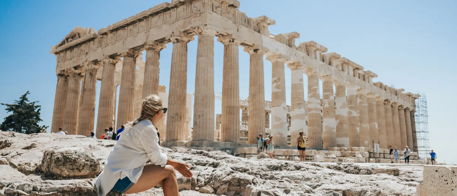 A person sitting on rocks views the Parthenon, an ancient temple with tall columns, under a clear blue sky.
