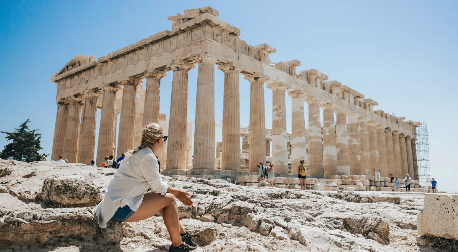 A person sitting on rocks views the Parthenon, an ancient temple with tall columns, under a clear blue sky.