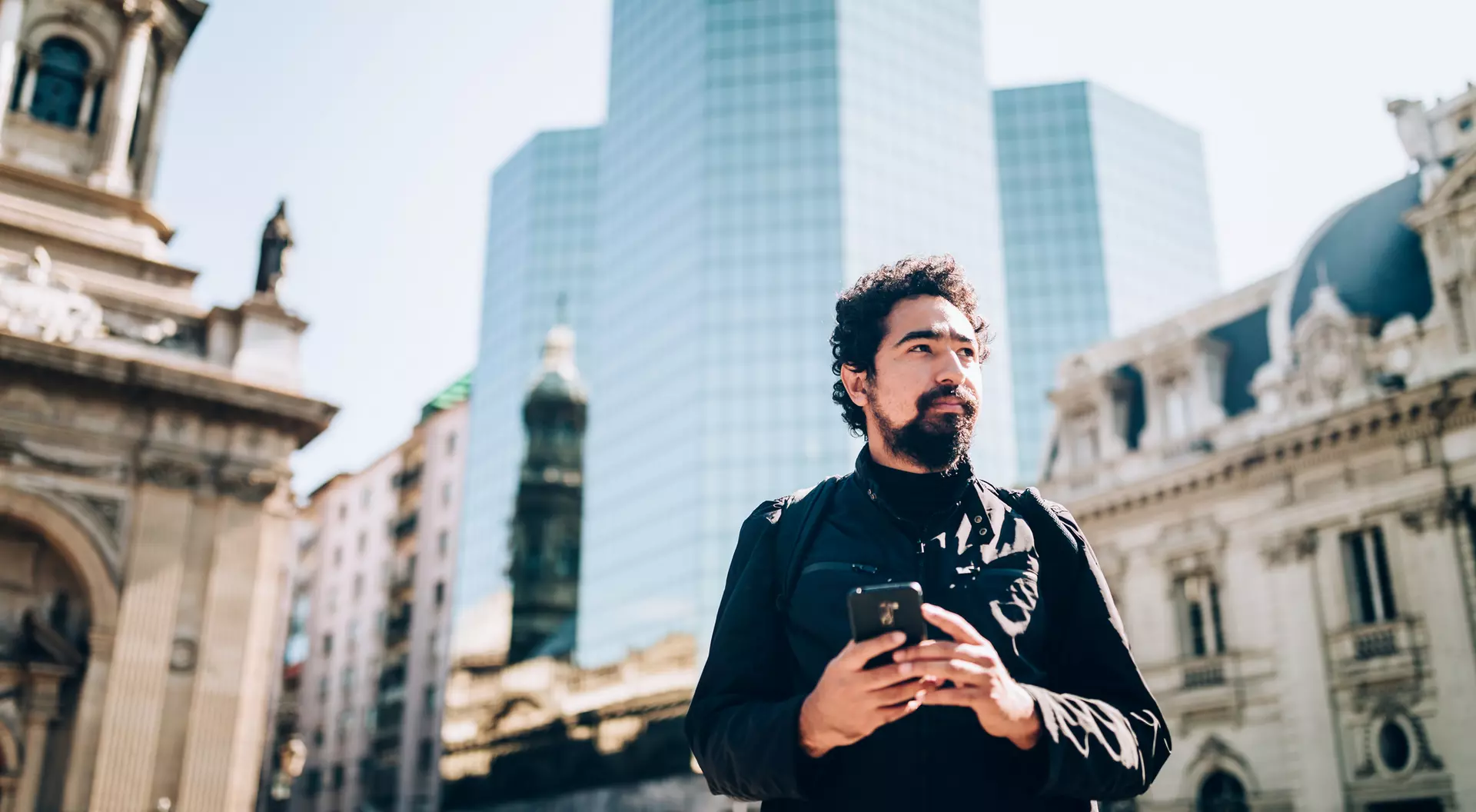 A man stands outdoors, holding a smartphone, with modern skyscrapers and historic buildings in the background.