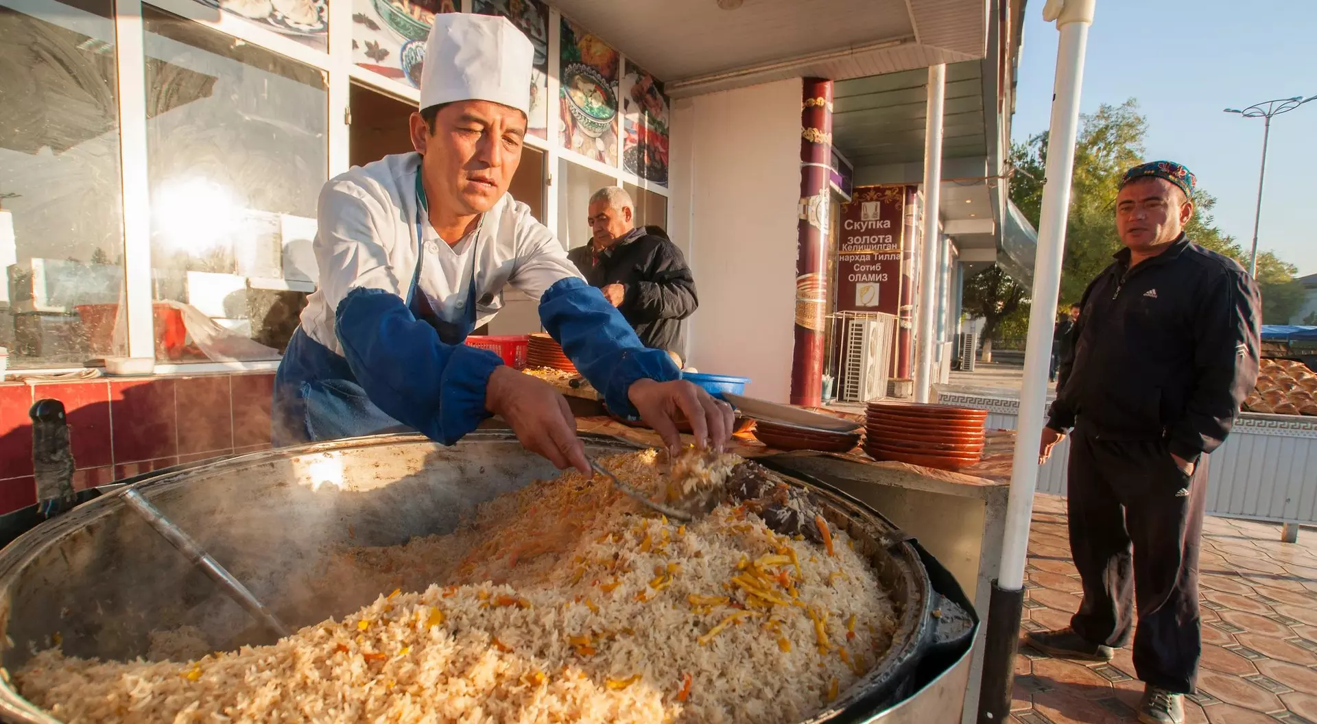 A chef in a white hat prepares a large pot of rice dish outdoors, while a man waits nearby.