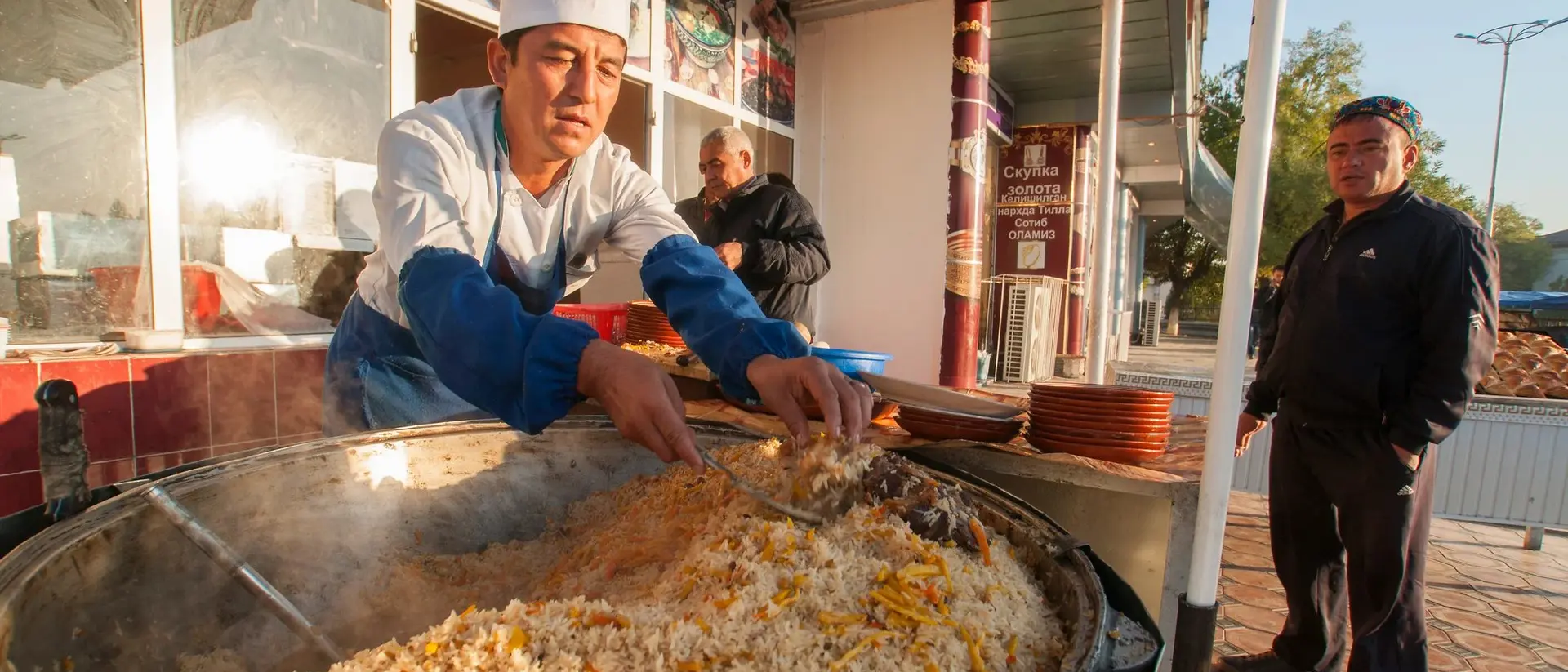 A chef in a white hat prepares a large pot of rice dish outdoors, while a man waits nearby.
