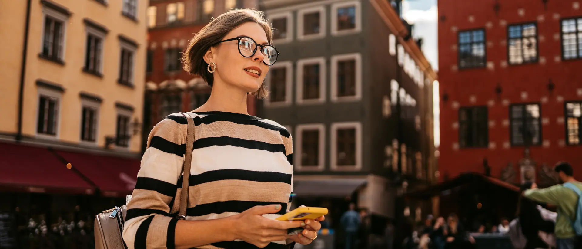 Young woman in a striped sweater stands in a plaza, holding a phone, with colorful buildings and people around her.