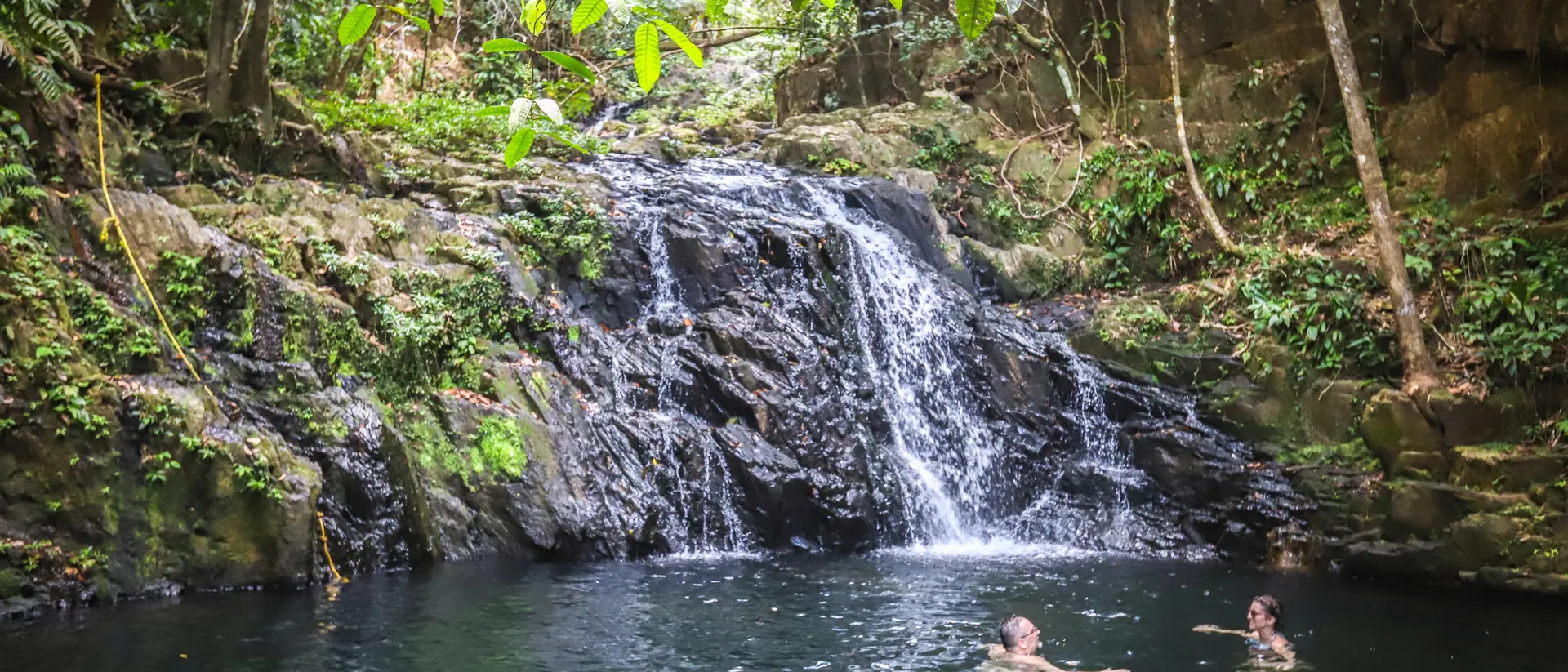 Two people swim in a natural pool beside a small waterfall, surrounded by lush green vegetation.
