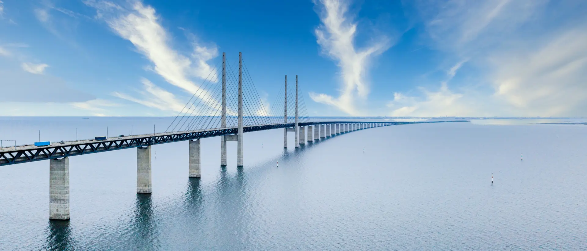 A long bridge spans over calm waters under a clear blue sky with wispy clouds.