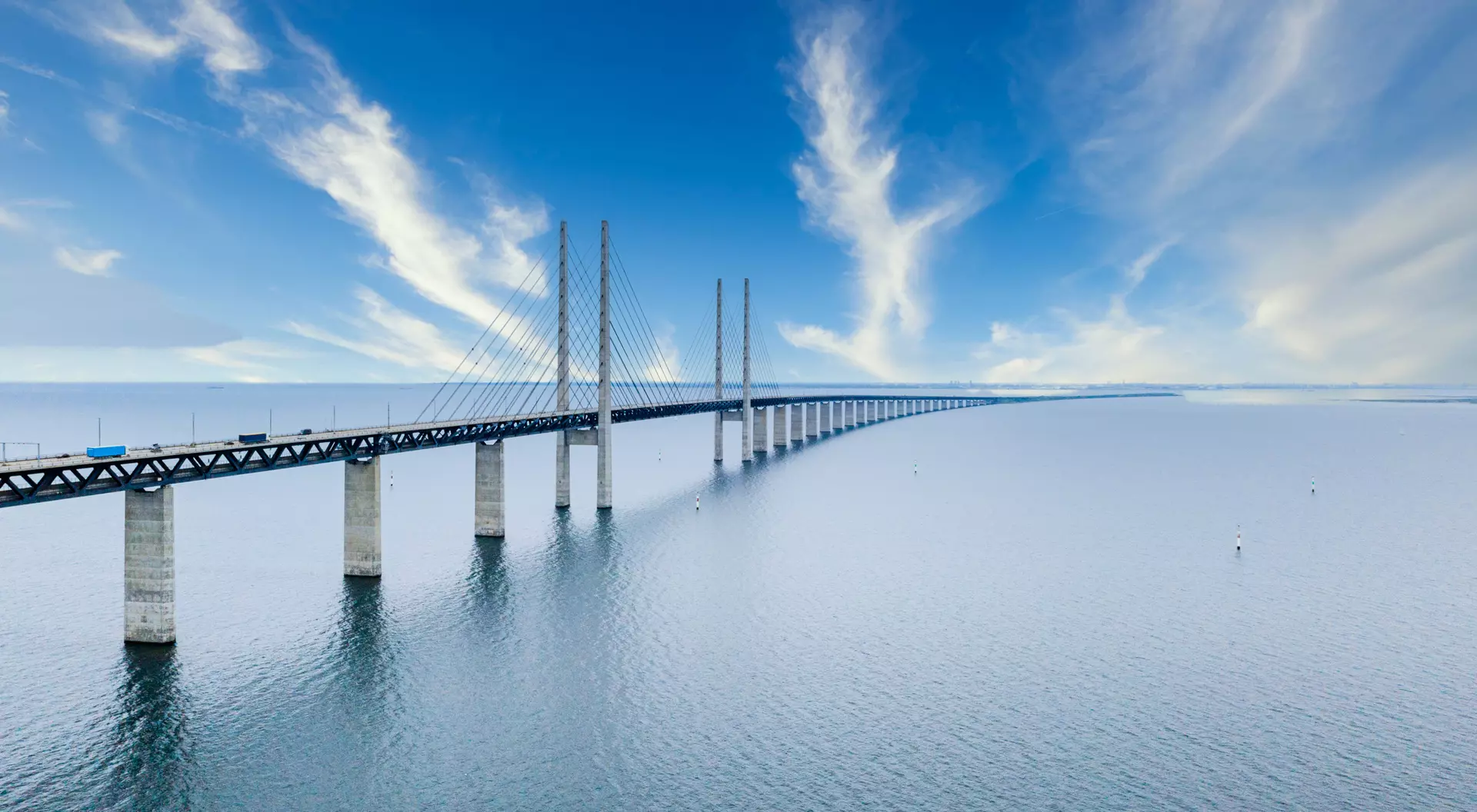 A long bridge spans over calm waters under a clear blue sky with wispy clouds.