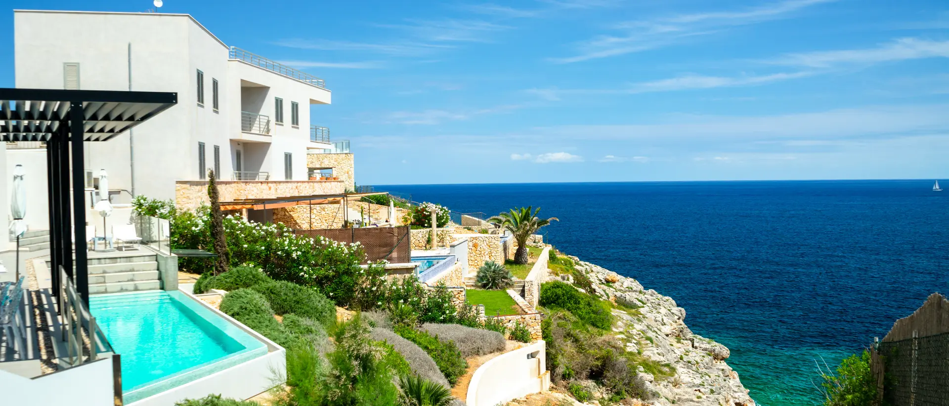 View of a coastal property with pools, gardens, and a rocky shoreline overlooking a blue ocean under a clear sky.