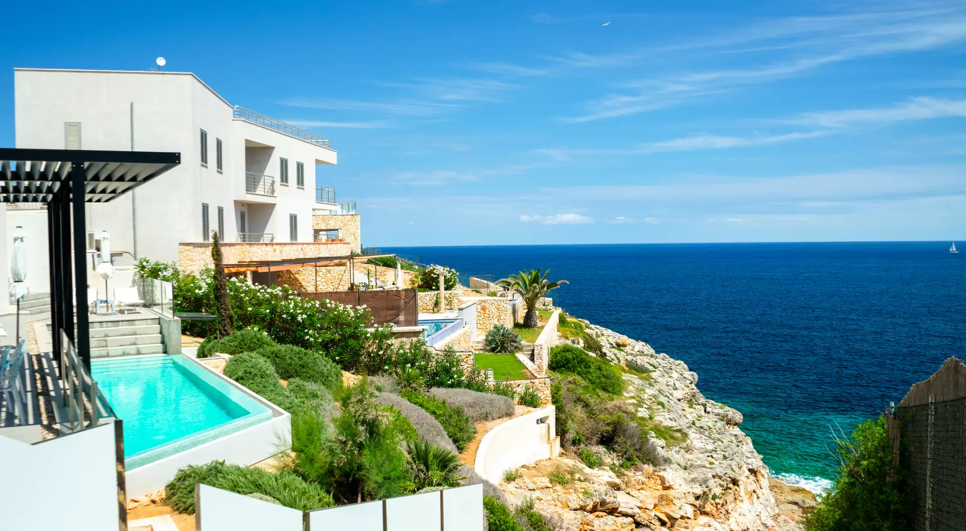 View of a coastal property with pools, gardens, and a rocky shoreline overlooking a blue ocean under a clear sky.