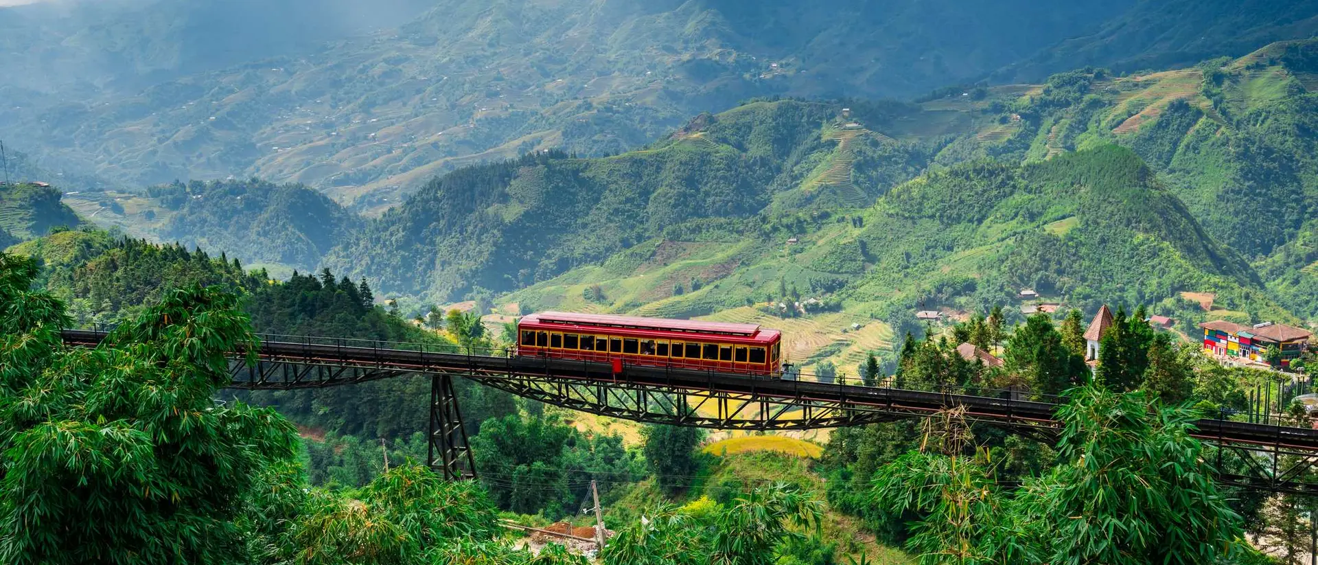 A red train crosses a bridge over lush green mountains under a cloudy sky.