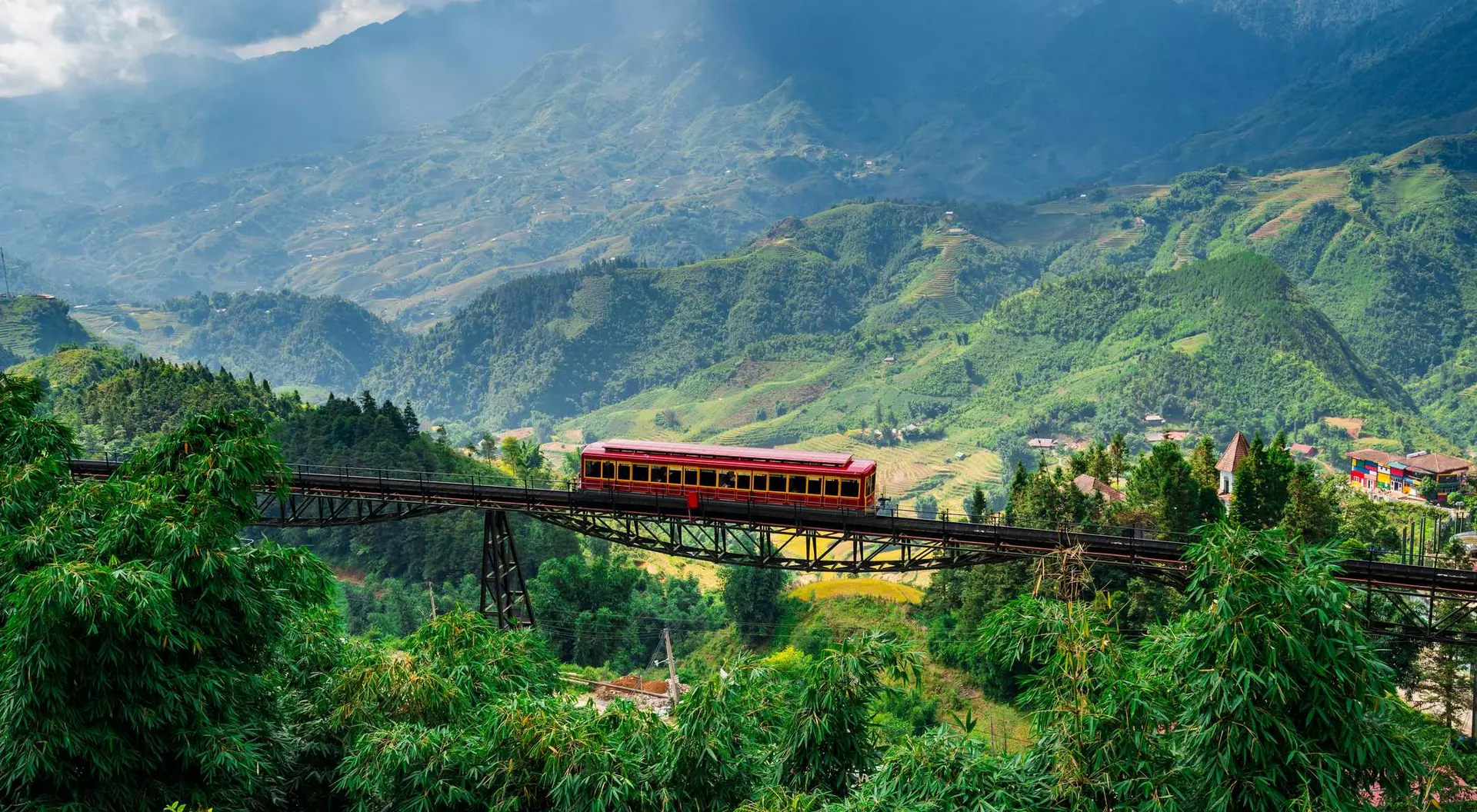 A red train crosses a bridge over lush green mountains under a cloudy sky.