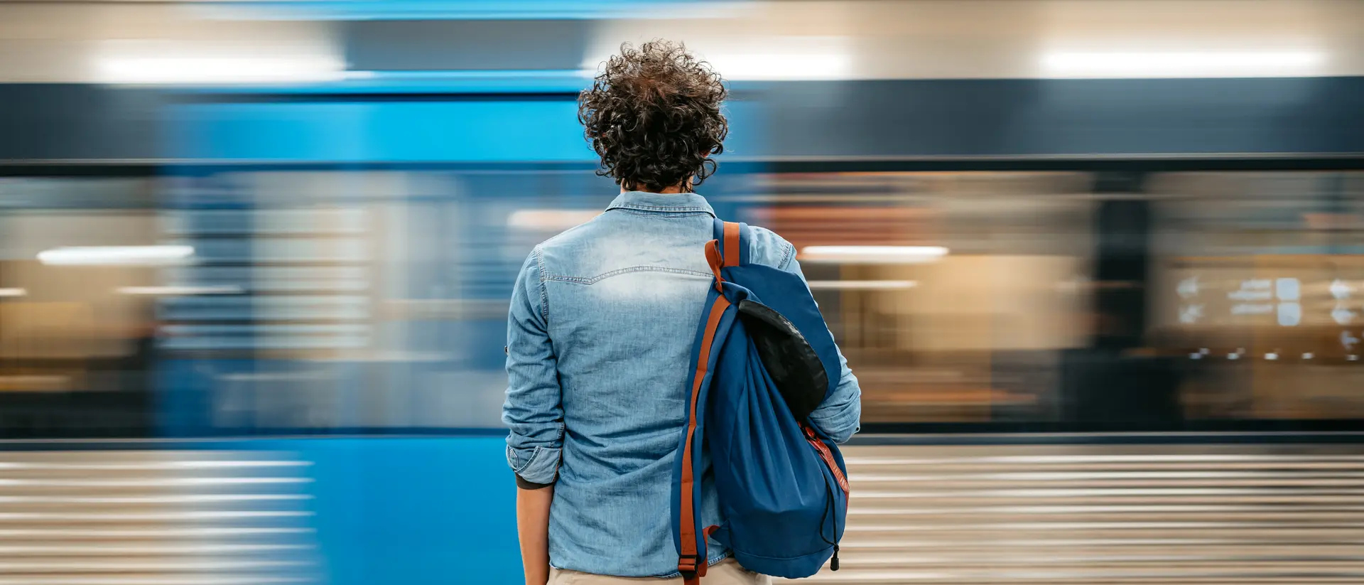 A person stands at a subway platform, facing a train speeding by, with a blue bag slung over their shoulder.
