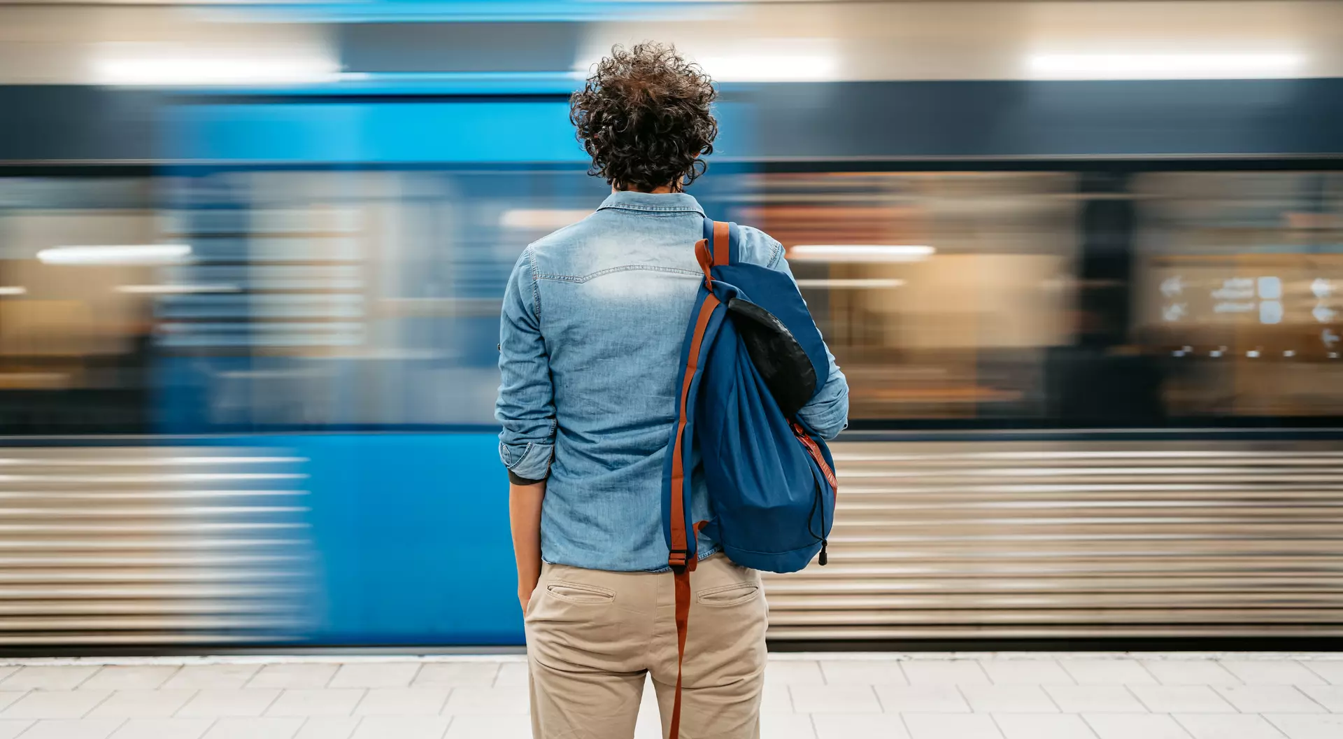 A person stands at a subway platform, facing a train speeding by, with a blue bag slung over their shoulder.
