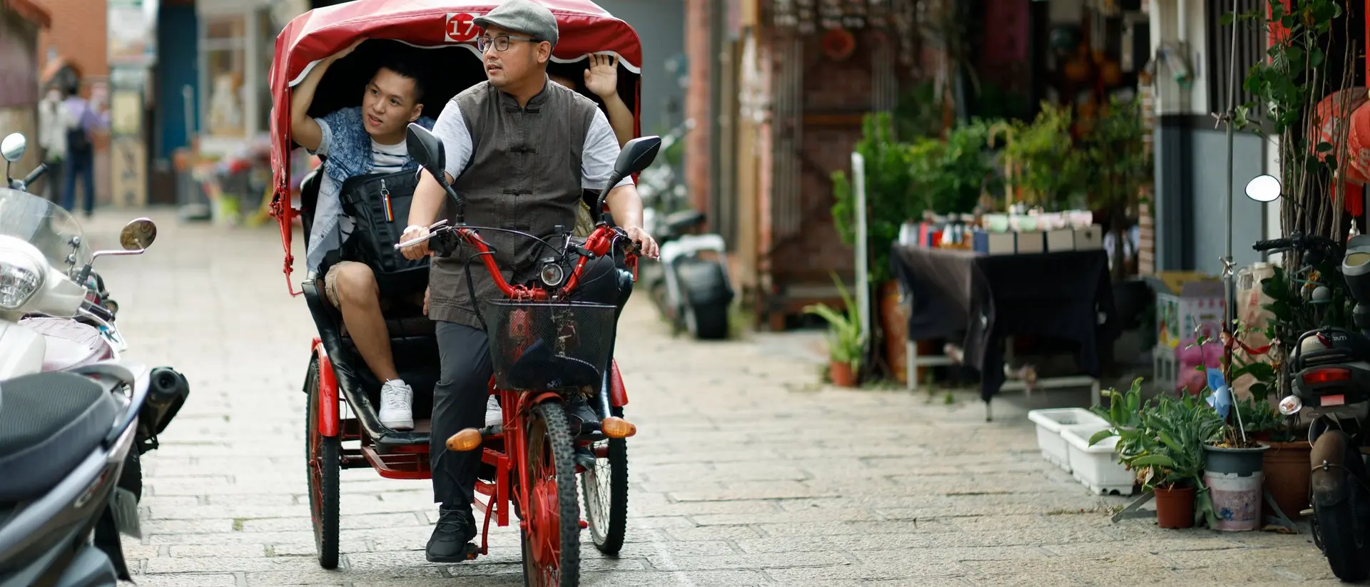 A man rides a pedicab with a child along a narrow street lined with shops and greenery.