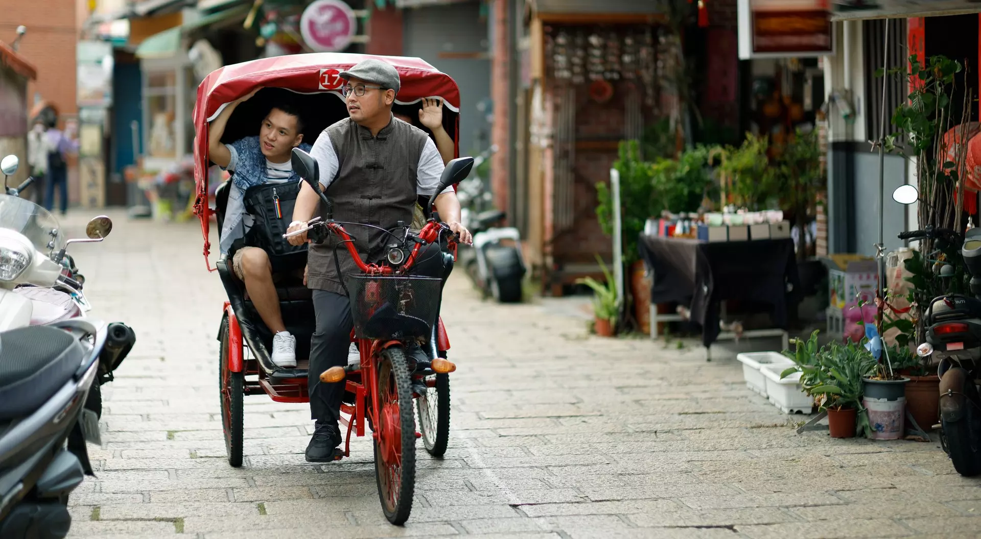 A man rides a pedicab with a child along a narrow street lined with shops and greenery.