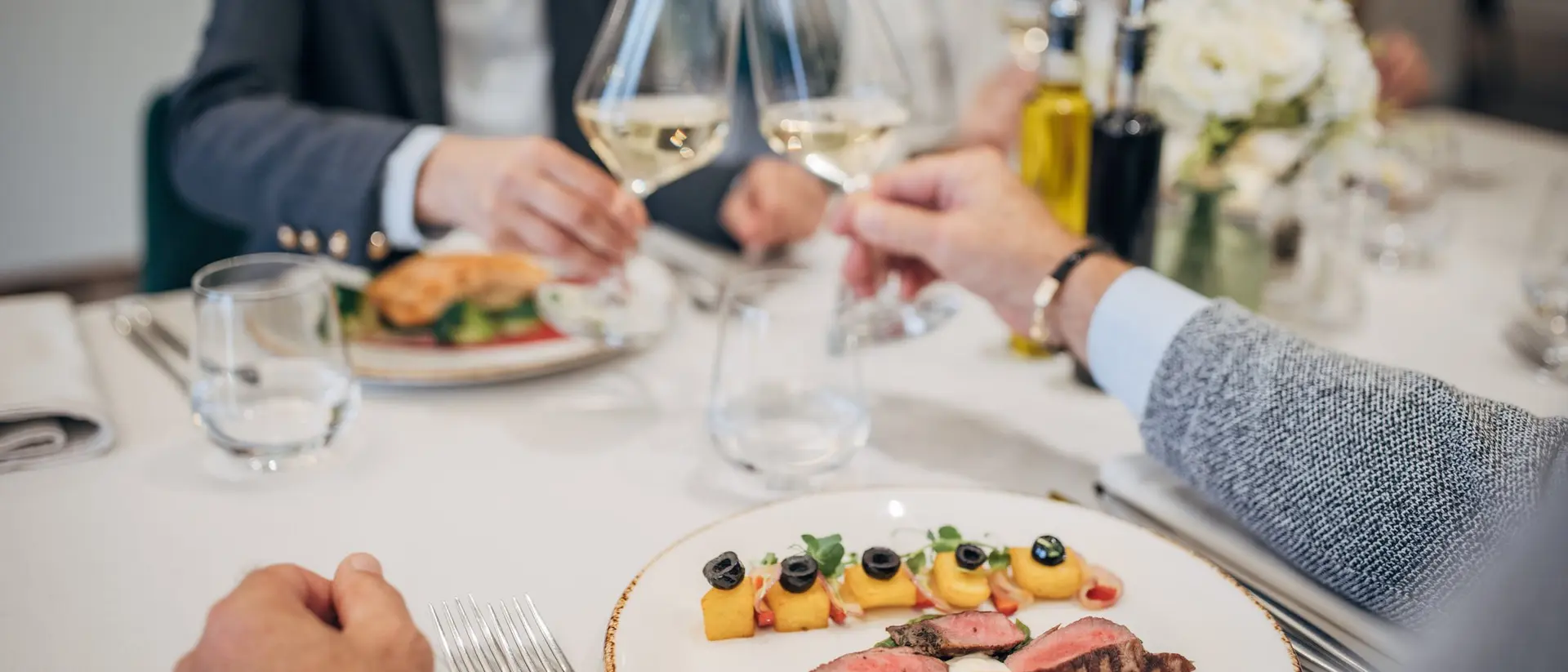 A dinner table set with a plated meal, wine glasses being toasted, and elegant dining decor.