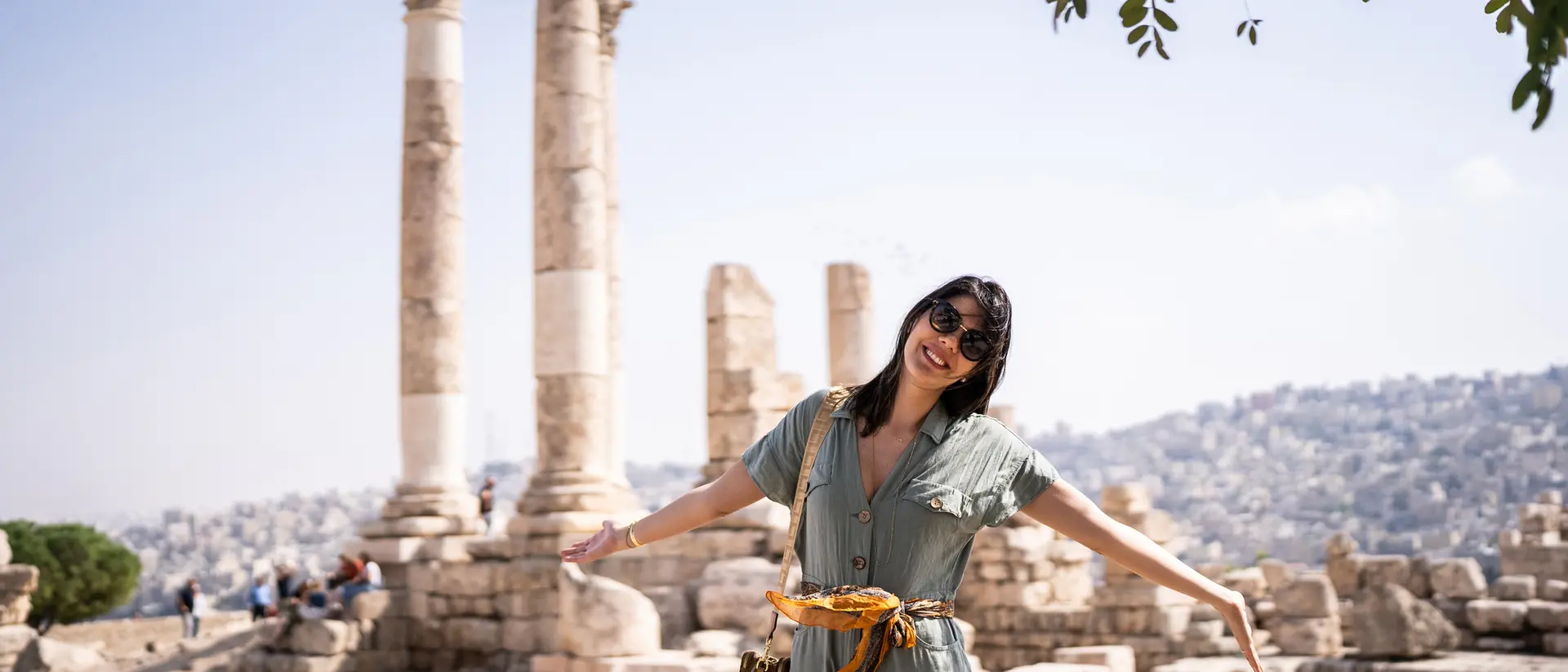 A woman in a green jumpsuit poses with arms outstretched in front of ancient pillars and a scenic background.