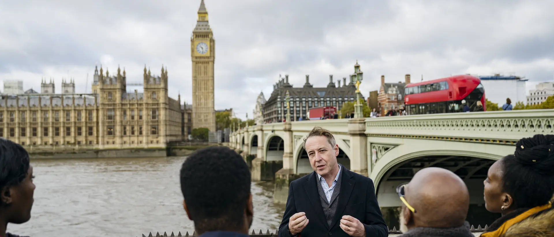 A tour guide speaks to a small group by the Thames, with Big Ben and the Houses of Parliament in the background.