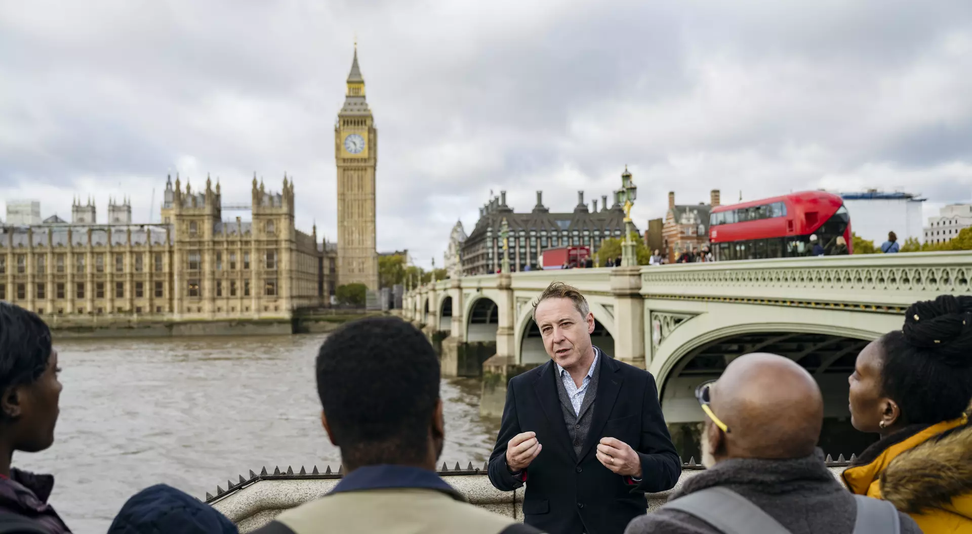 A tour guide speaks to a small group by the Thames, with Big Ben and the Houses of Parliament in the background.
