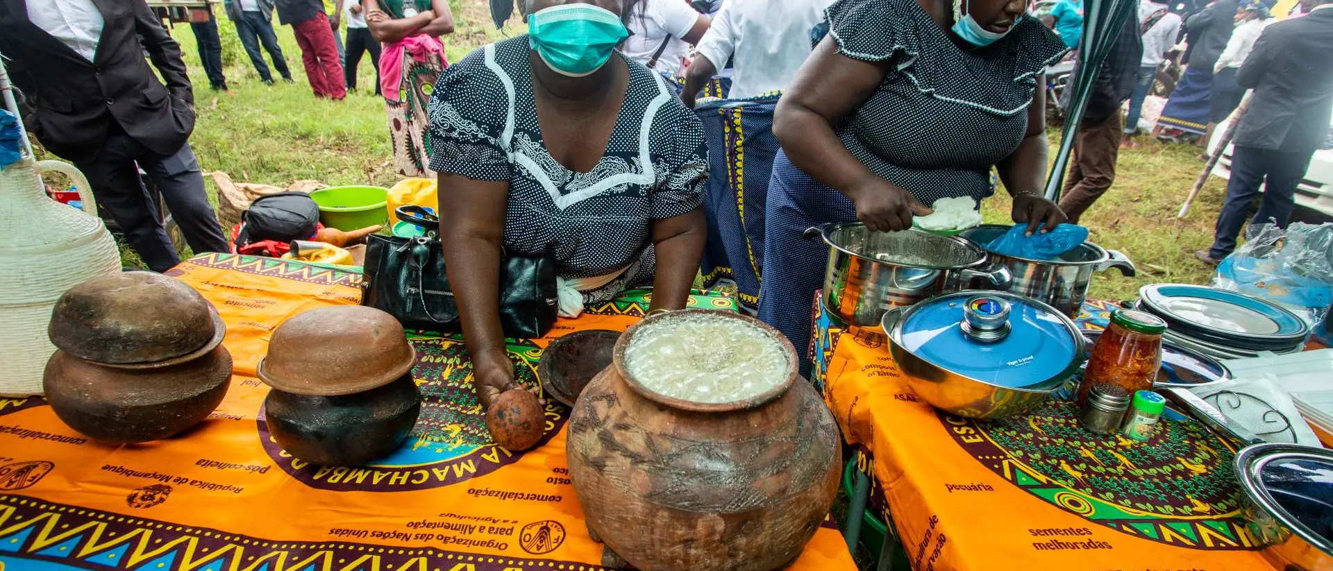 Two women prepare food outdoors at a vibrant market, with traditional dishes displayed on colorful patterned tablecloths.