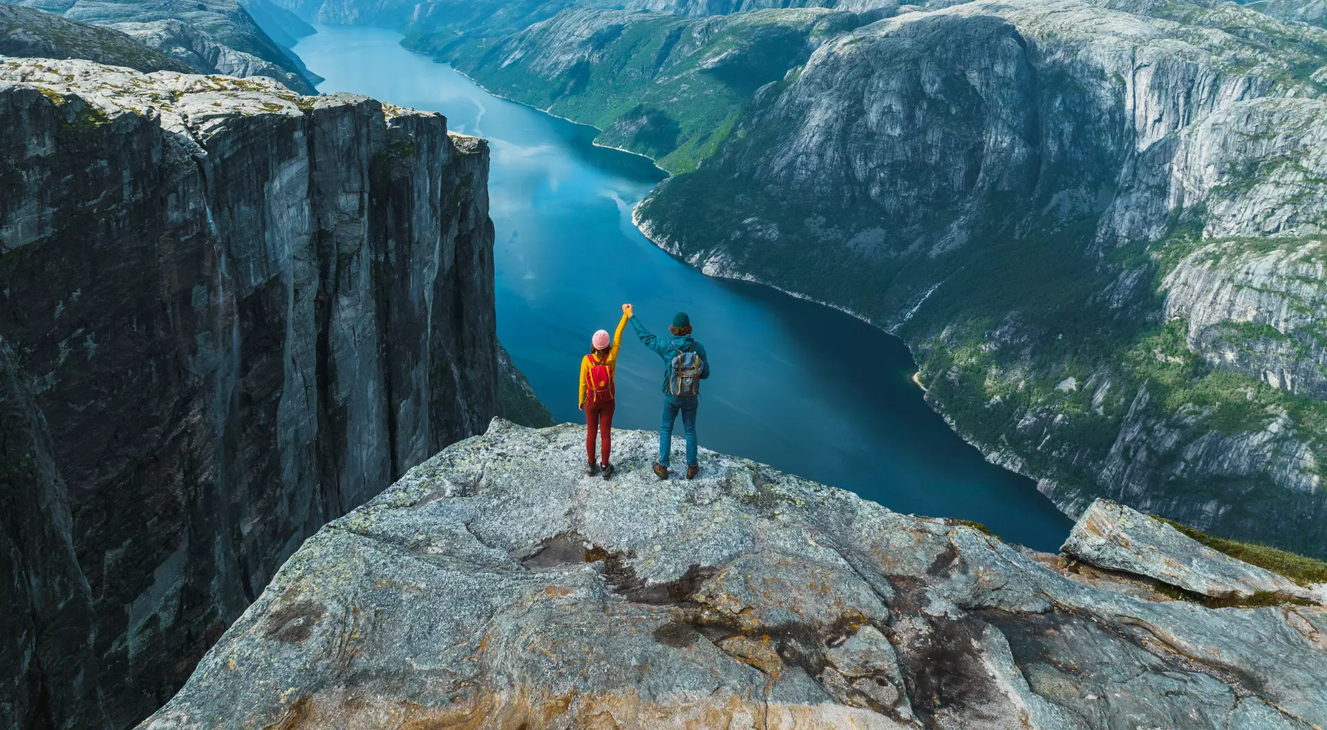 Two hikers stand on a rocky ledge, overlooking a deep blue fjord surrounded by lush green mountains and a cloudy sky.