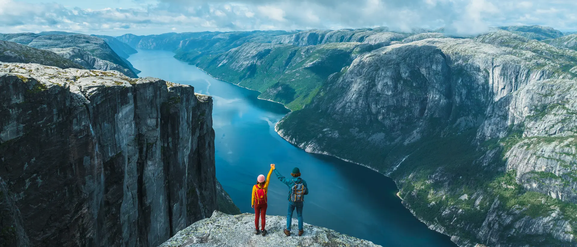 Two hikers stand on a rocky ledge, overlooking a deep blue fjord surrounded by lush green mountains and a cloudy sky.