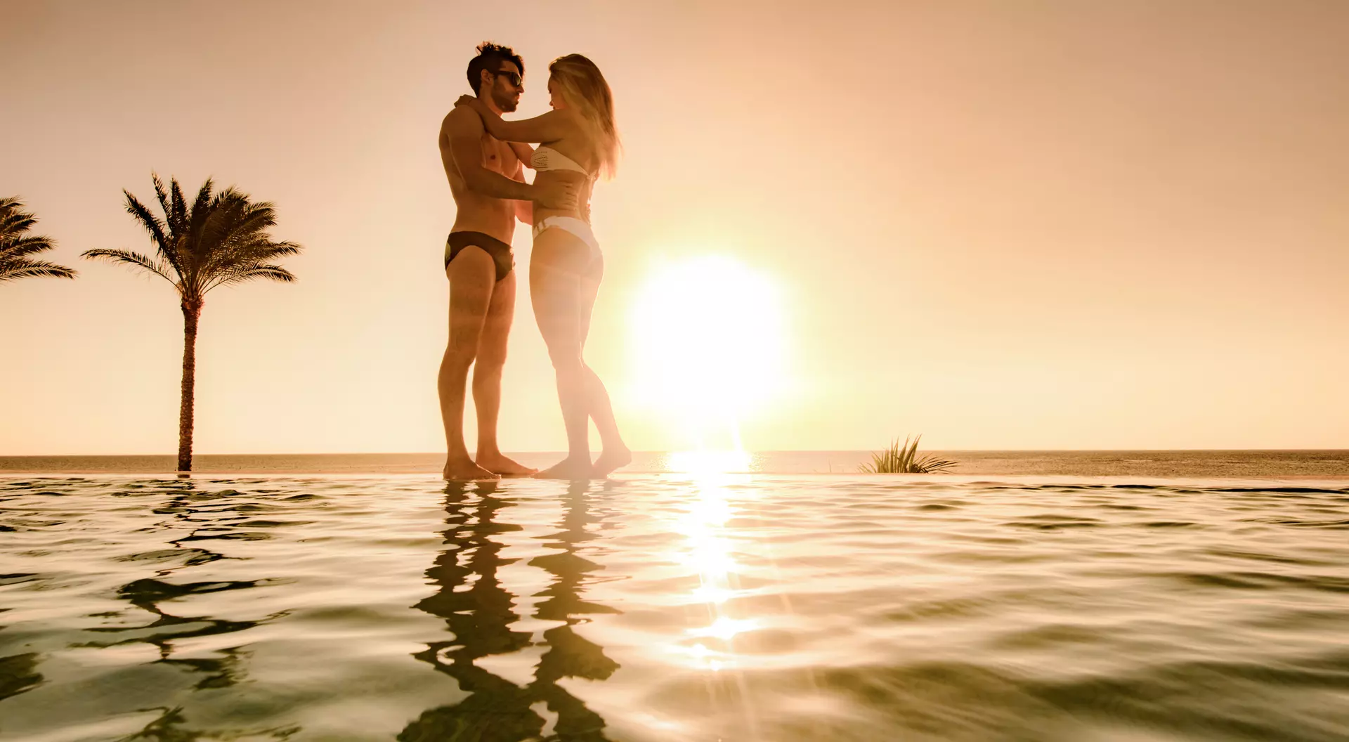 A couple embraces in an infinity pool at sunset, surrounded by palm trees.