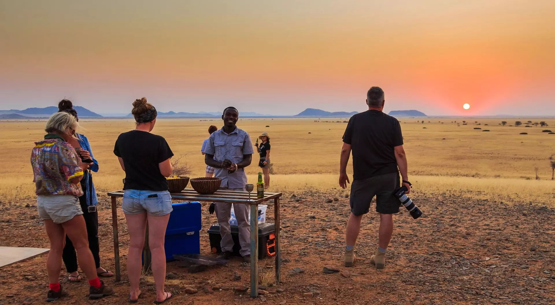 People gather around a table in a sunset-lit desert, enjoying a meal while a photographer captures the moment.