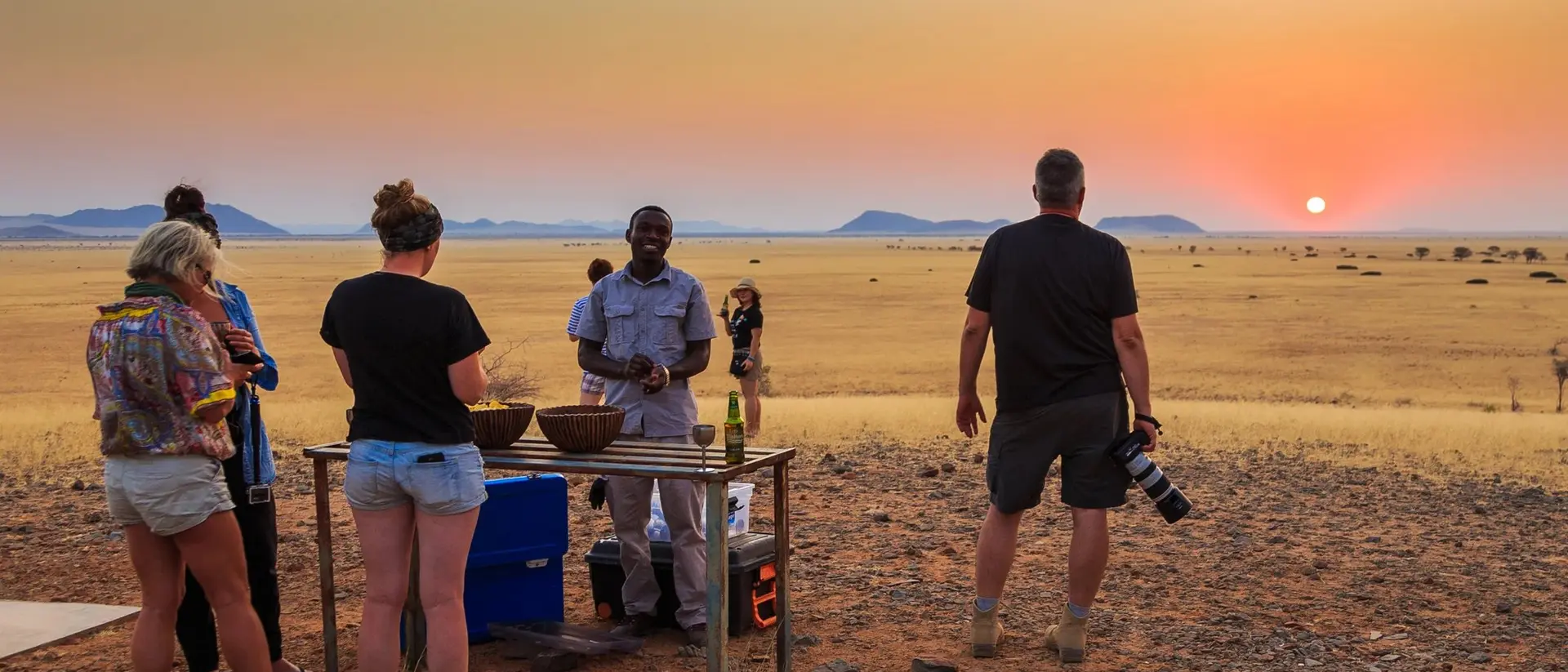 People gather around a table in a sunset-lit desert, enjoying a meal while a photographer captures the moment.