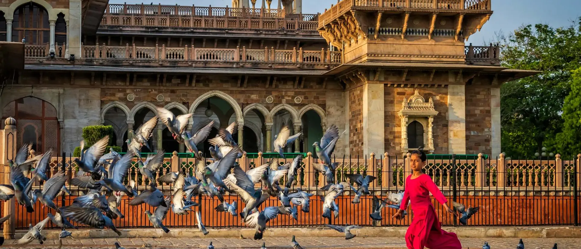 Person in a red outfit runs joyfully amid a flock of pigeons in front of a historic building at sunrise.