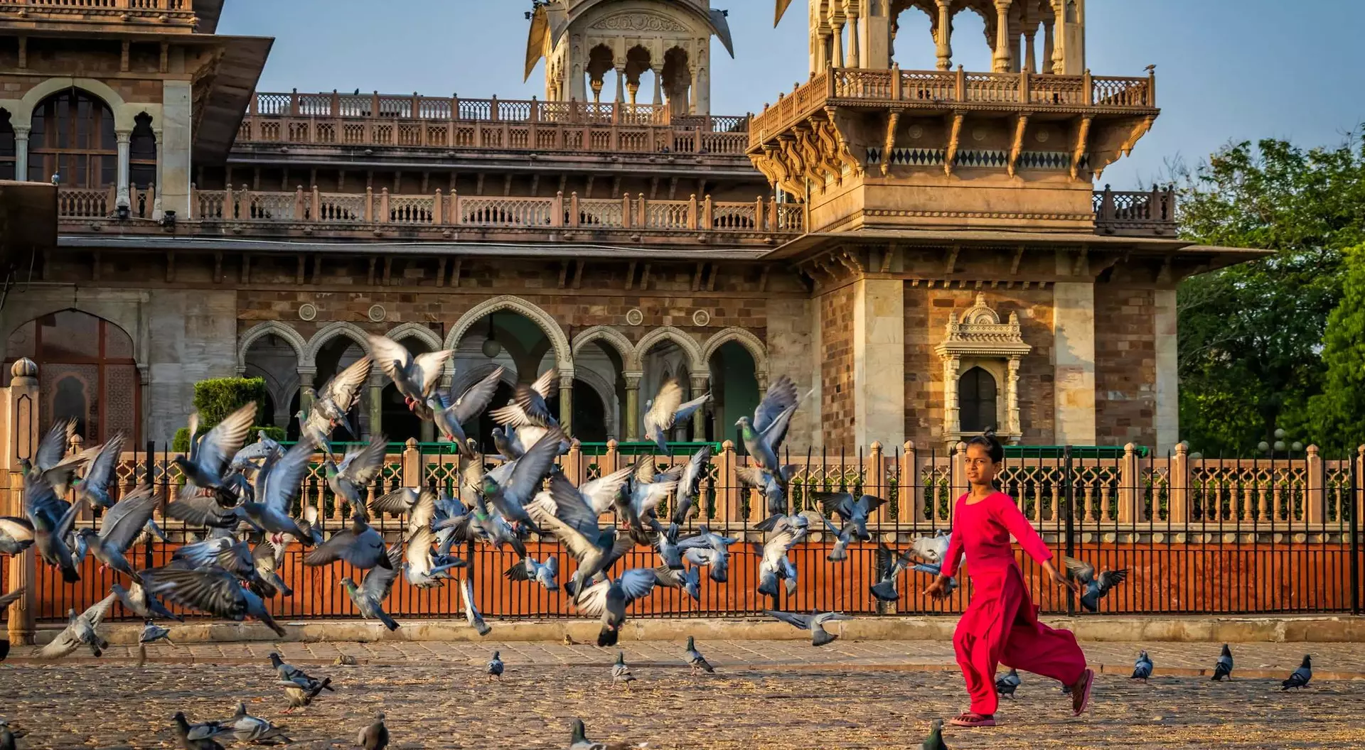 Person in a red outfit runs joyfully amid a flock of pigeons in front of a historic building at sunrise.