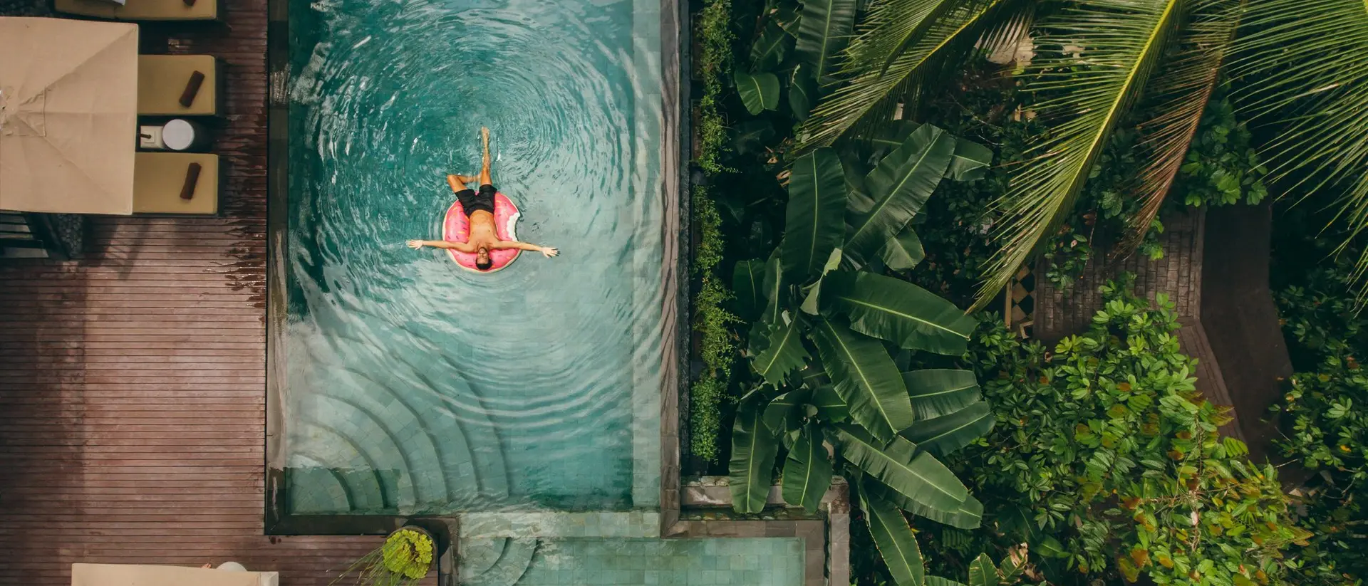 Aerial view of a person floating in a pool surrounded by lush greenery and lounge areas.