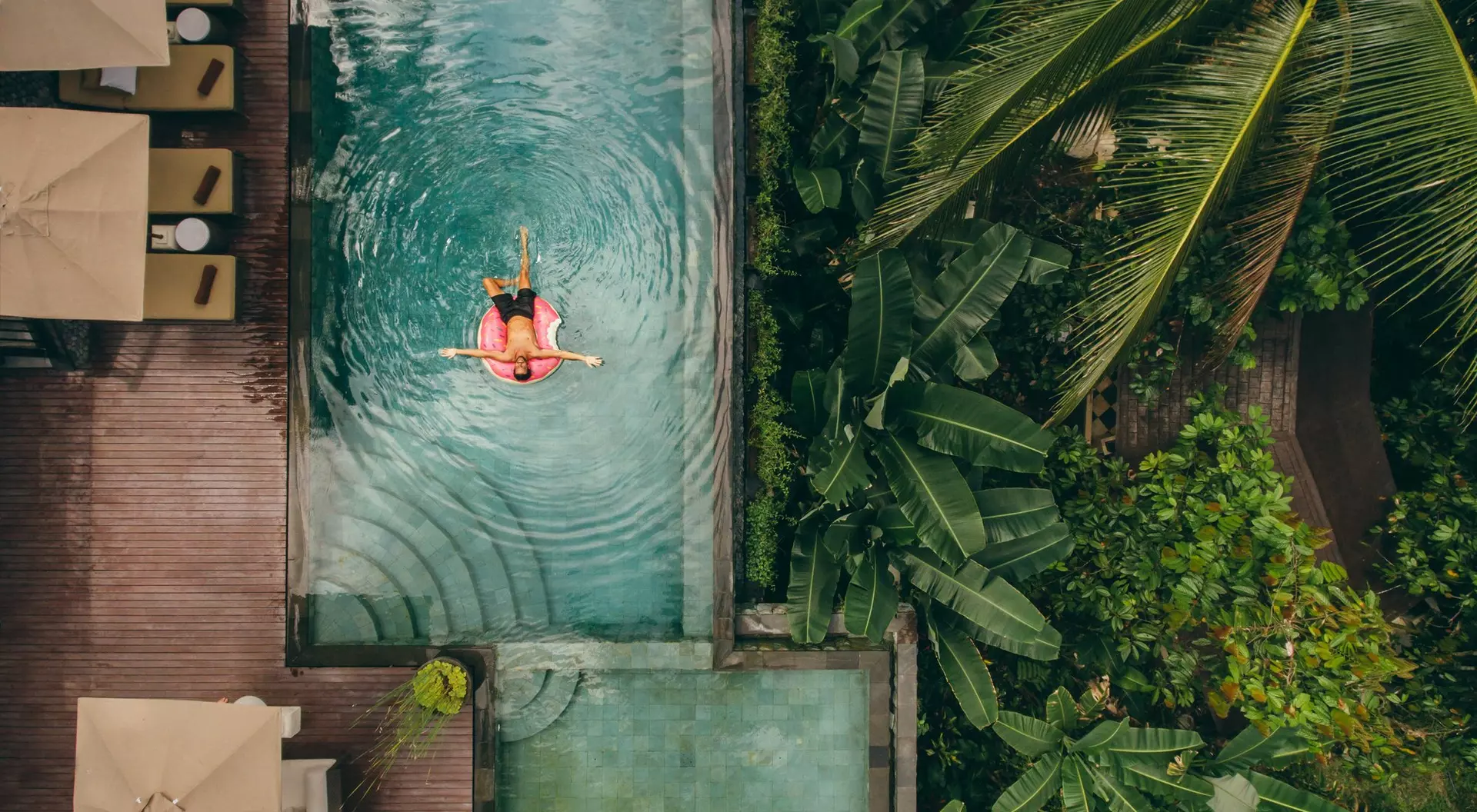Aerial view of a person floating in a pool surrounded by lush greenery and lounge areas.