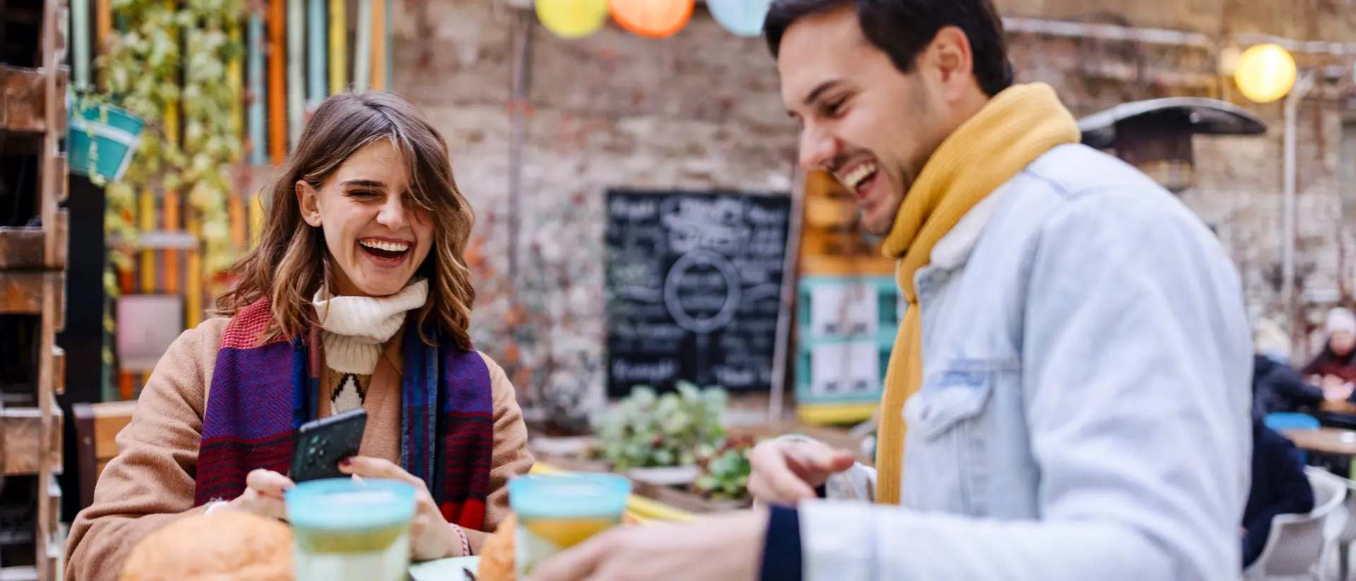 A man and woman sit at a colorful outdoor cafe, laughing and enjoying drinks and pastries.